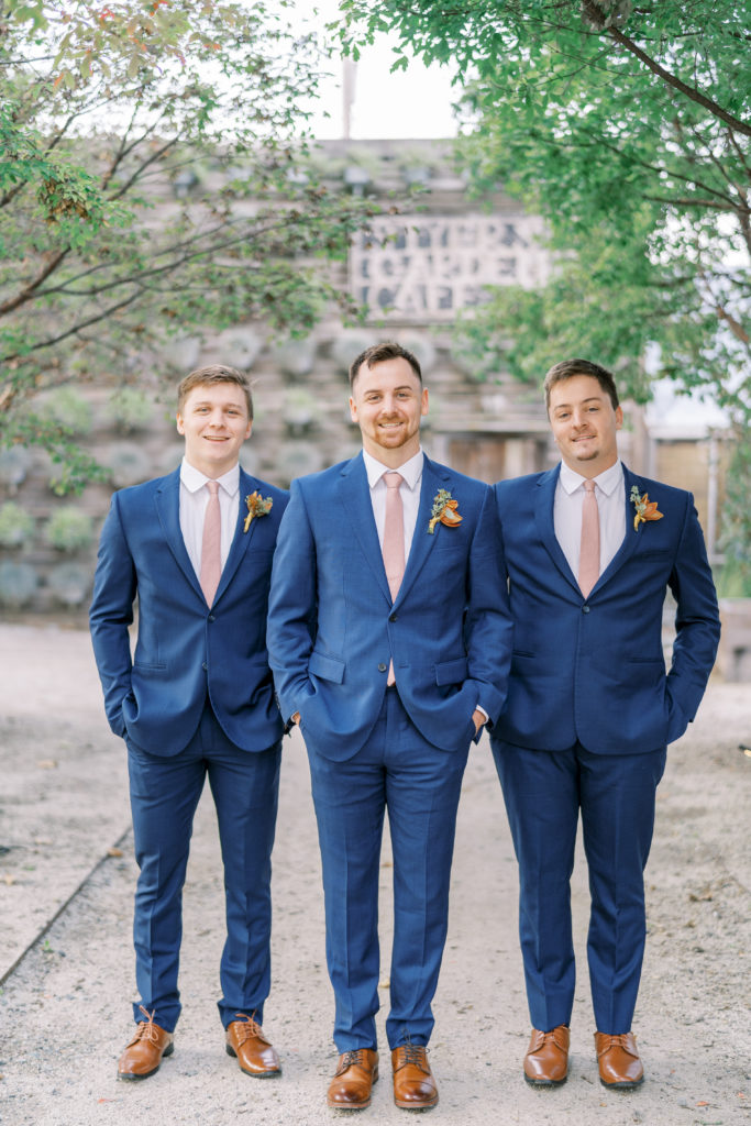 Groom and groomsmen pose on gravel pathway with blue suit and pink tie 