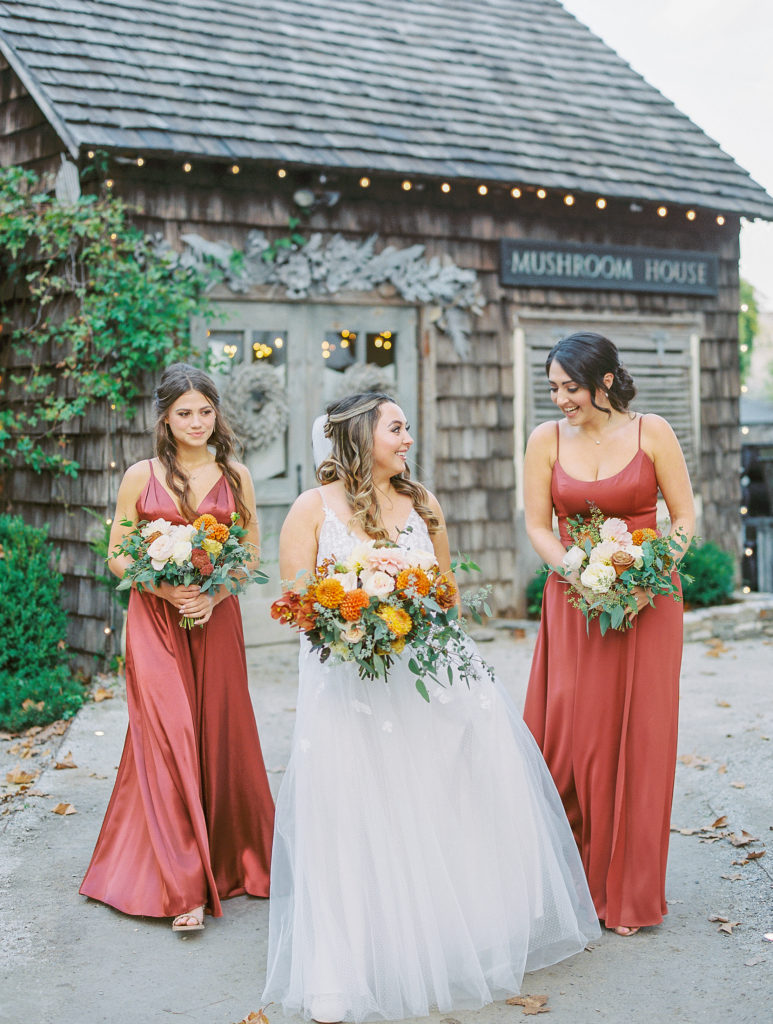 Bride and bridesmaids walk along pathway in orange-red dresses 