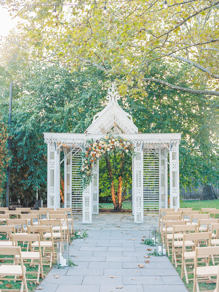 White pavilion with autumnal colored flowers along arch and stone pathway aisle for Terrain at Styer's Wedding
