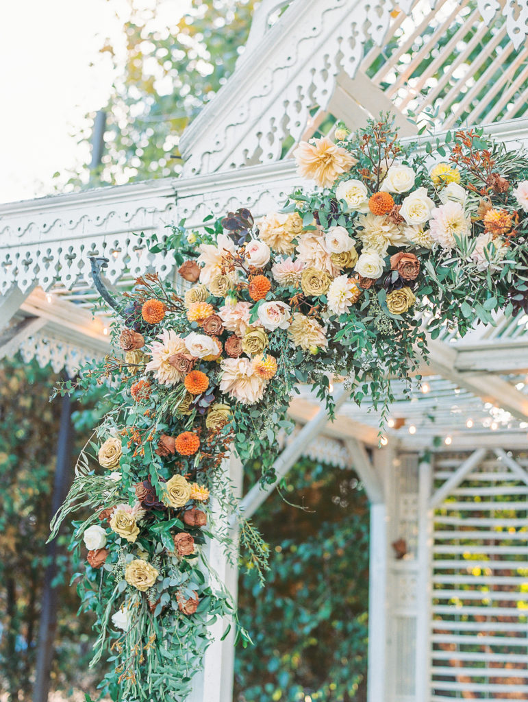 Side view of white antique pavilion with cream, yellow, pink, and orange flowers hanging off arch 