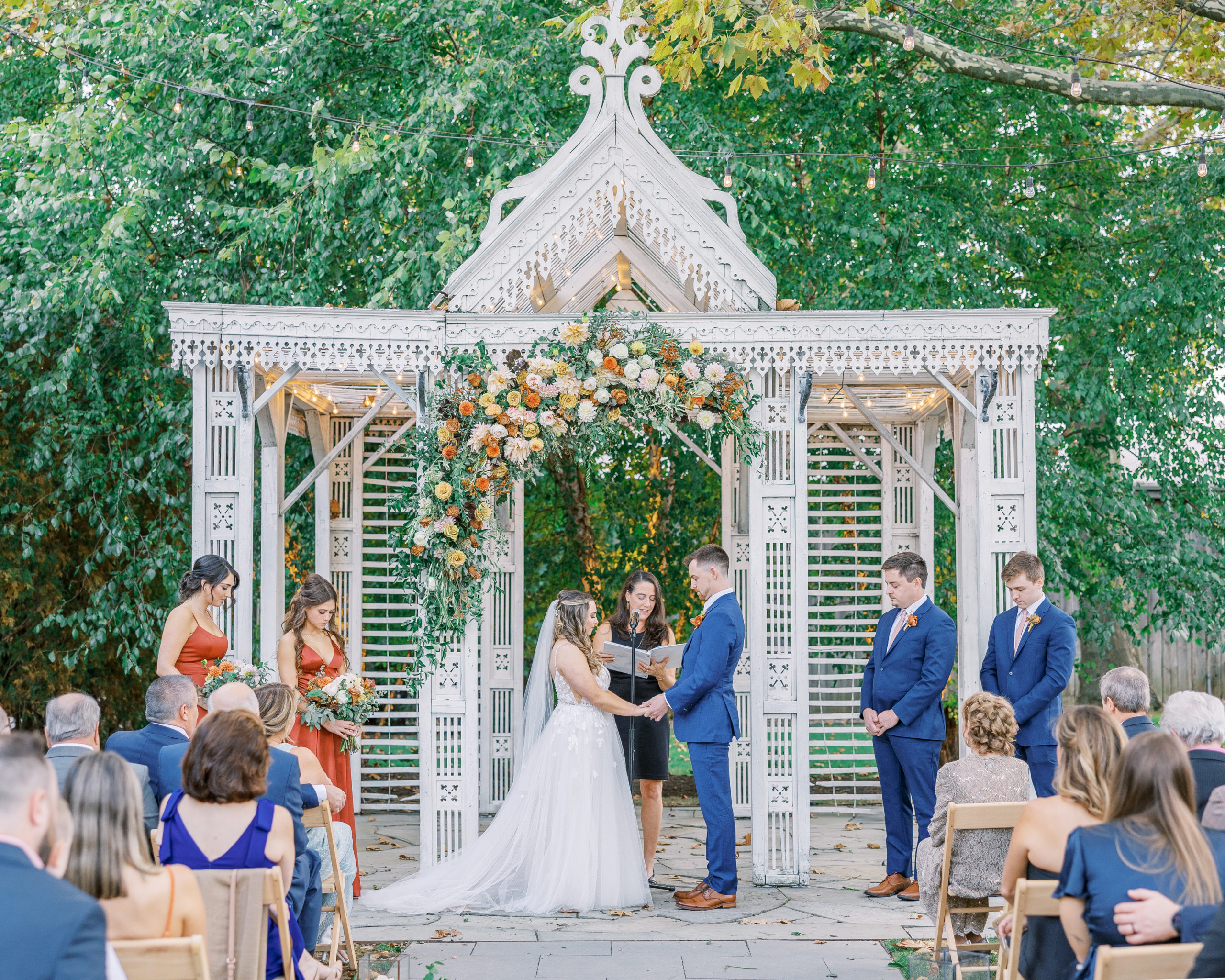 Bride and groom hold hands at altar with white pavilion and autumnal flowers cascading down for Terrain at Styer's Wedding