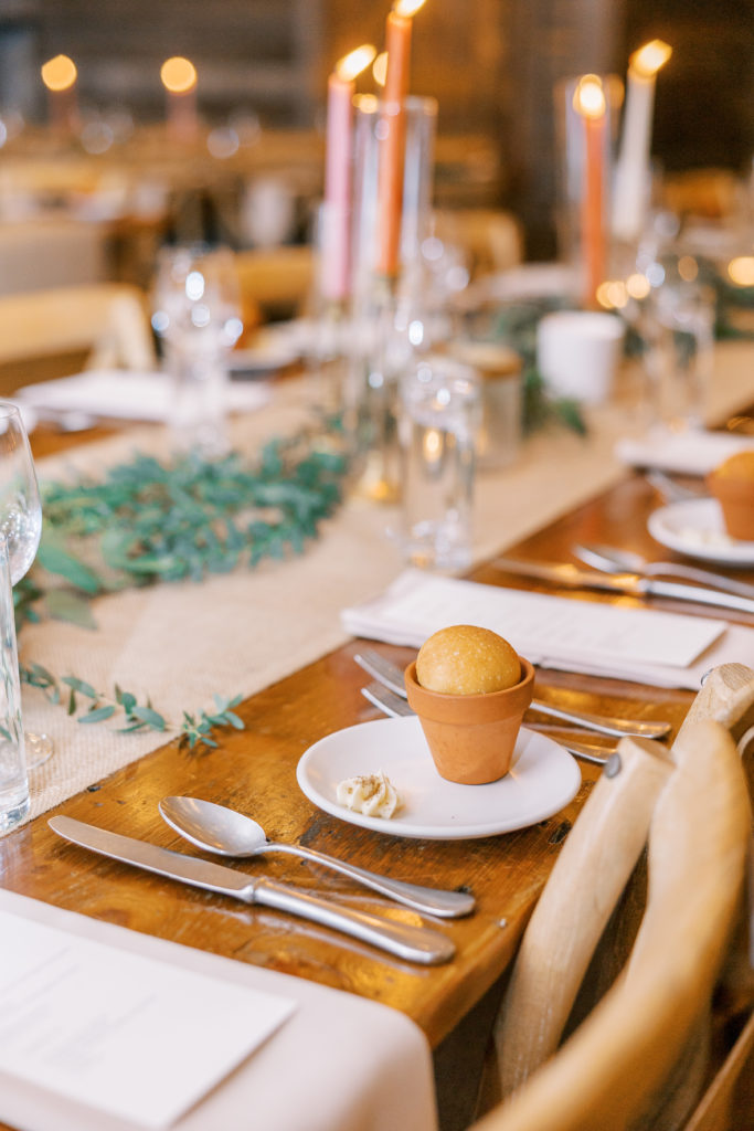 Wedding reception table with silverware, tall pink candles, and small bread in garden pot 