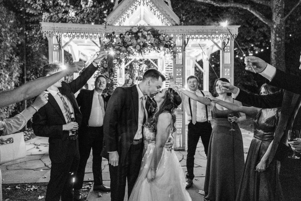 Bride and groom share a kiss for their sendoff as guests hold sparklers over them with white pavilion behind them for Terrain Glen Mills Wedding
