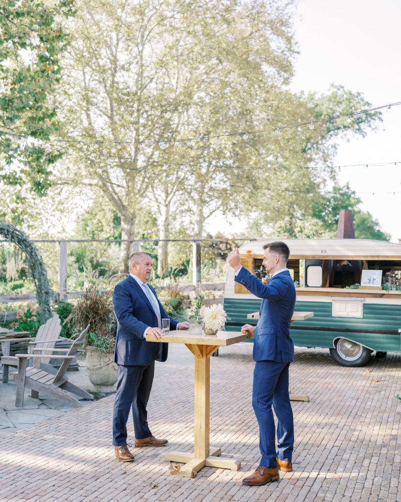 Groom and father share a drink at bar table outside in blue suits for Terrain Glen Mills Wedding