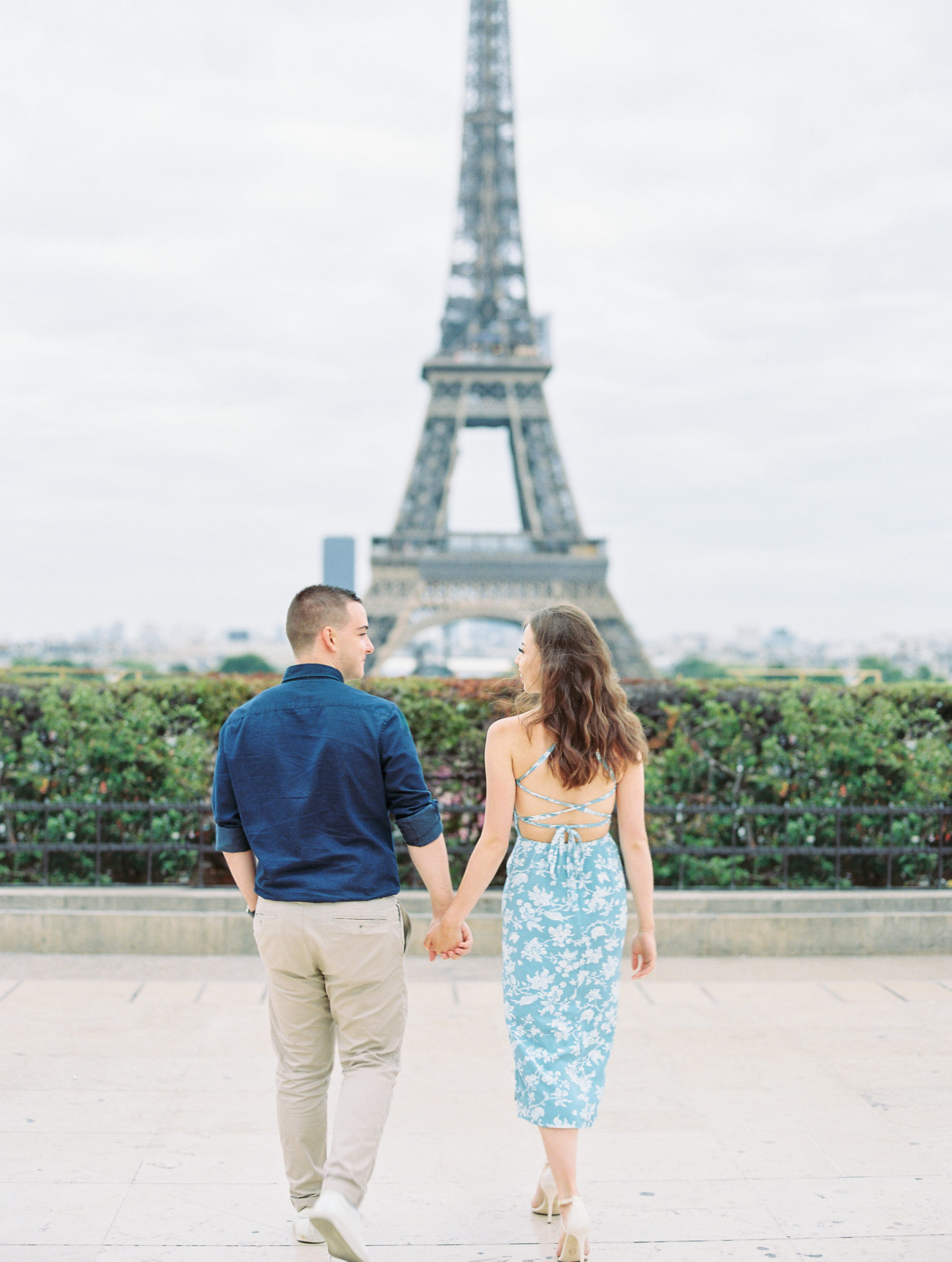 Couple smiles and holds hands walking towards Eiffel Tower on stone pathway for Paris Film Photographer