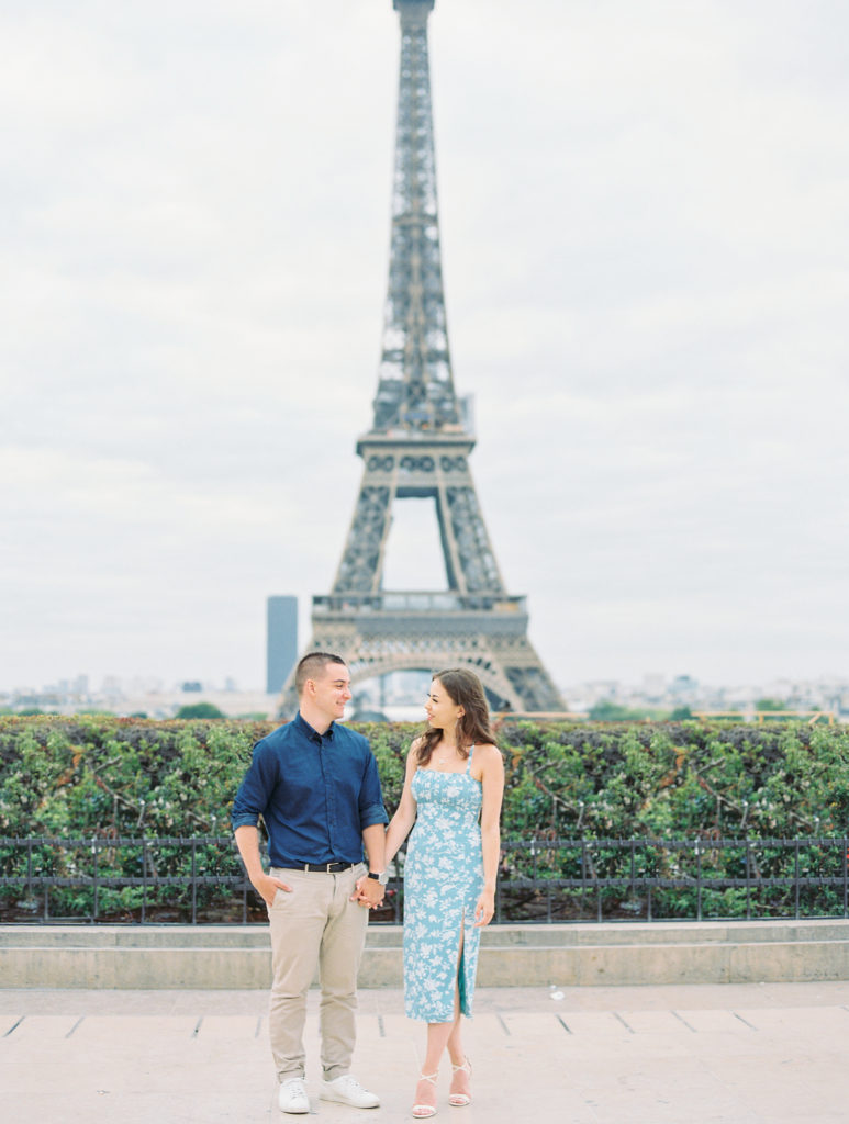Couple holds hands and smiles in front of Eiffel Tower for Paris Engagement Session