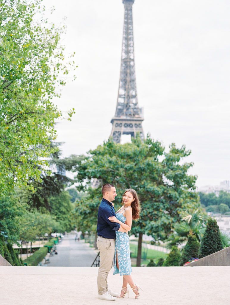 Couple embrace and smile in front of Eiffel Tower with greenery and trees for Paris Engagement Session