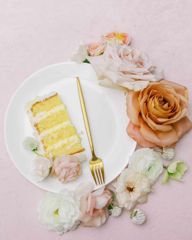 Piece of vanilla cake on plate with gold fork and peach and white flowers around plate 