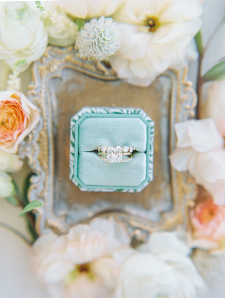 Green ring box with wedding ring on small gold tray surrounded by pink and cream flowers 