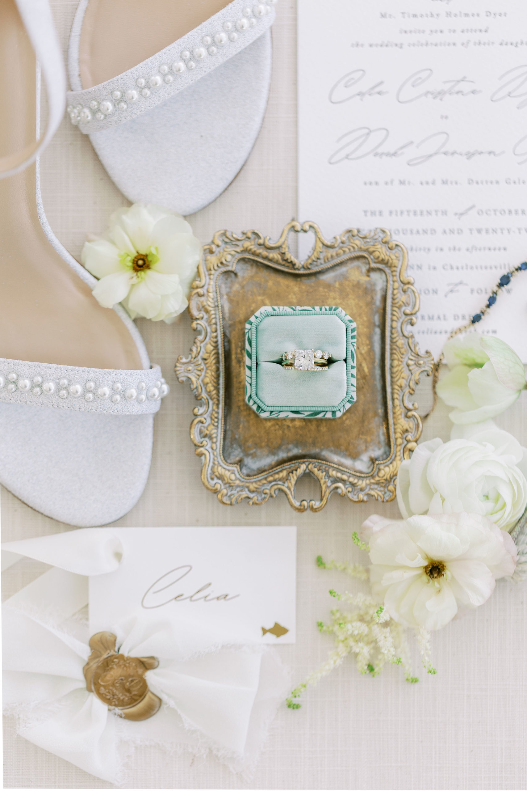 Flat lay of cream wedding invitations, white high heels with small pearls, cream flowers, and wedding ring in green box on gold tray for Clifton wedding photography