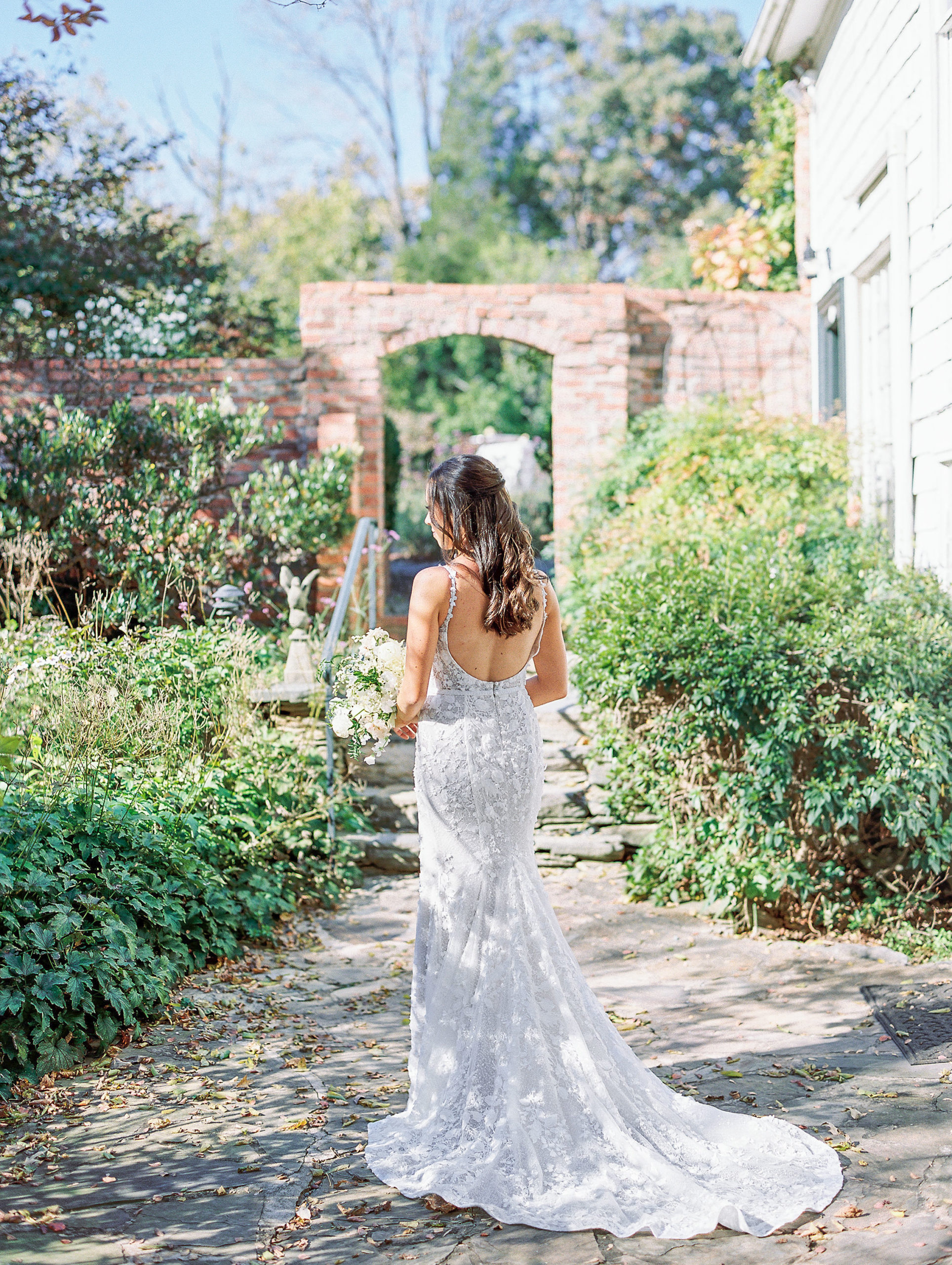 Bride is turned away in garden holding bouquet with brick arch behind for Clifton wedding photography