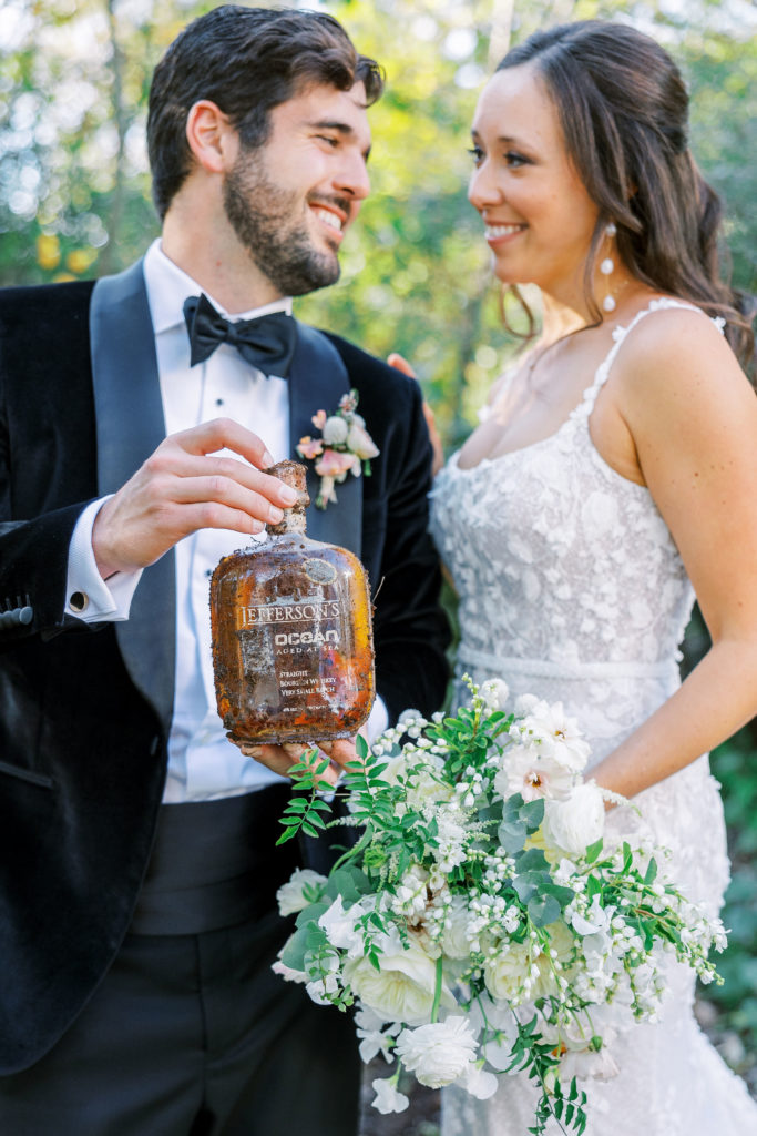 Bride and groom smile and hold up whiskey bottle 