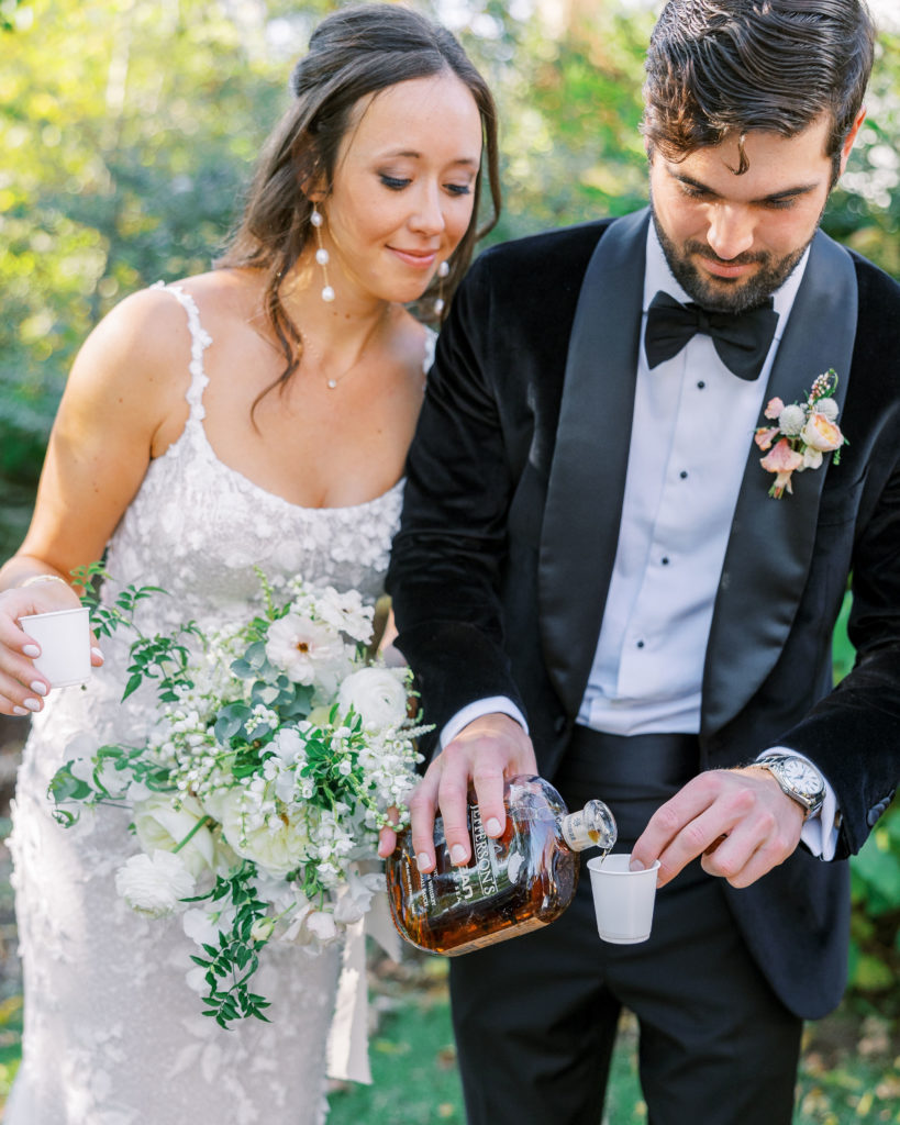 Groom pours whiskey into a cup and bride leans over and watches, holding bouquet for Clifton wedding photography