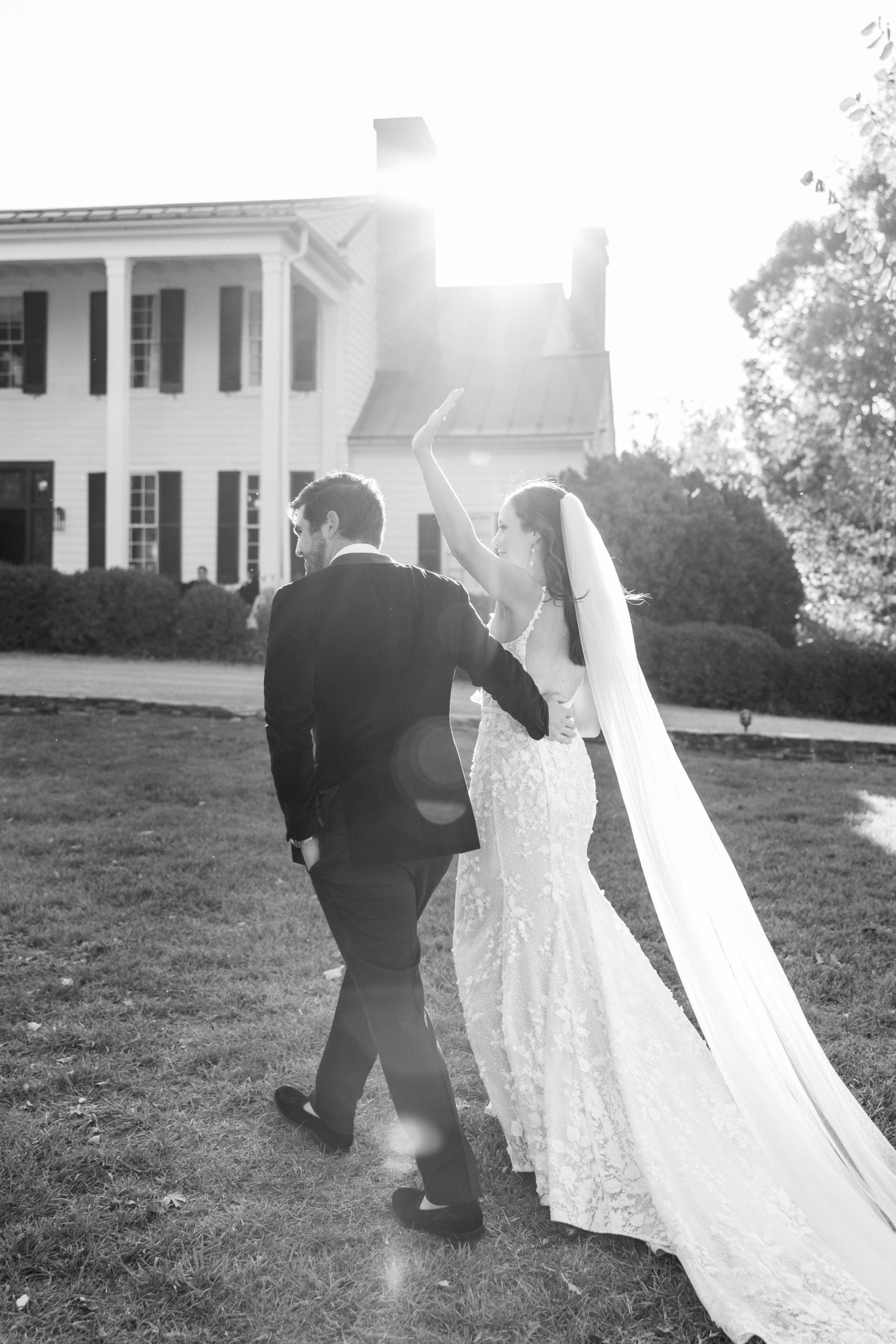 Bride waves and groom has hand on her back as they walk across lawn in front of white house for Clifton wedding photography