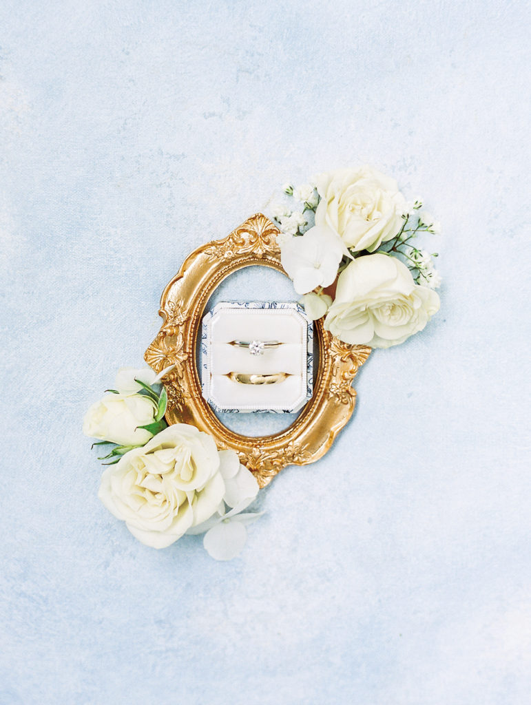 Wedding rings with golden frame and white roses on light blue surface 