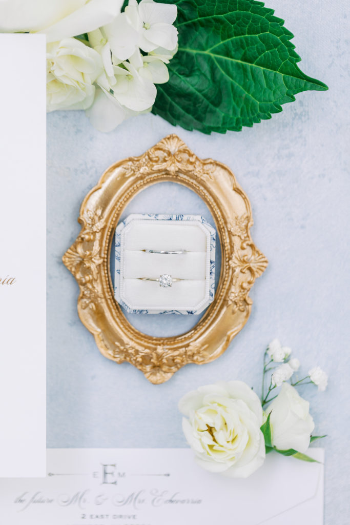 Wedding rings in white and blue ring box with gold frame around it with sides of ivory invitations and white roses 