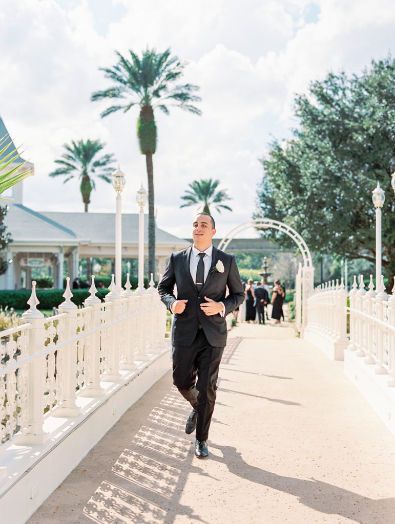 Groom walks over white bridge wearing black suit for Disney Film Wedding Photographer