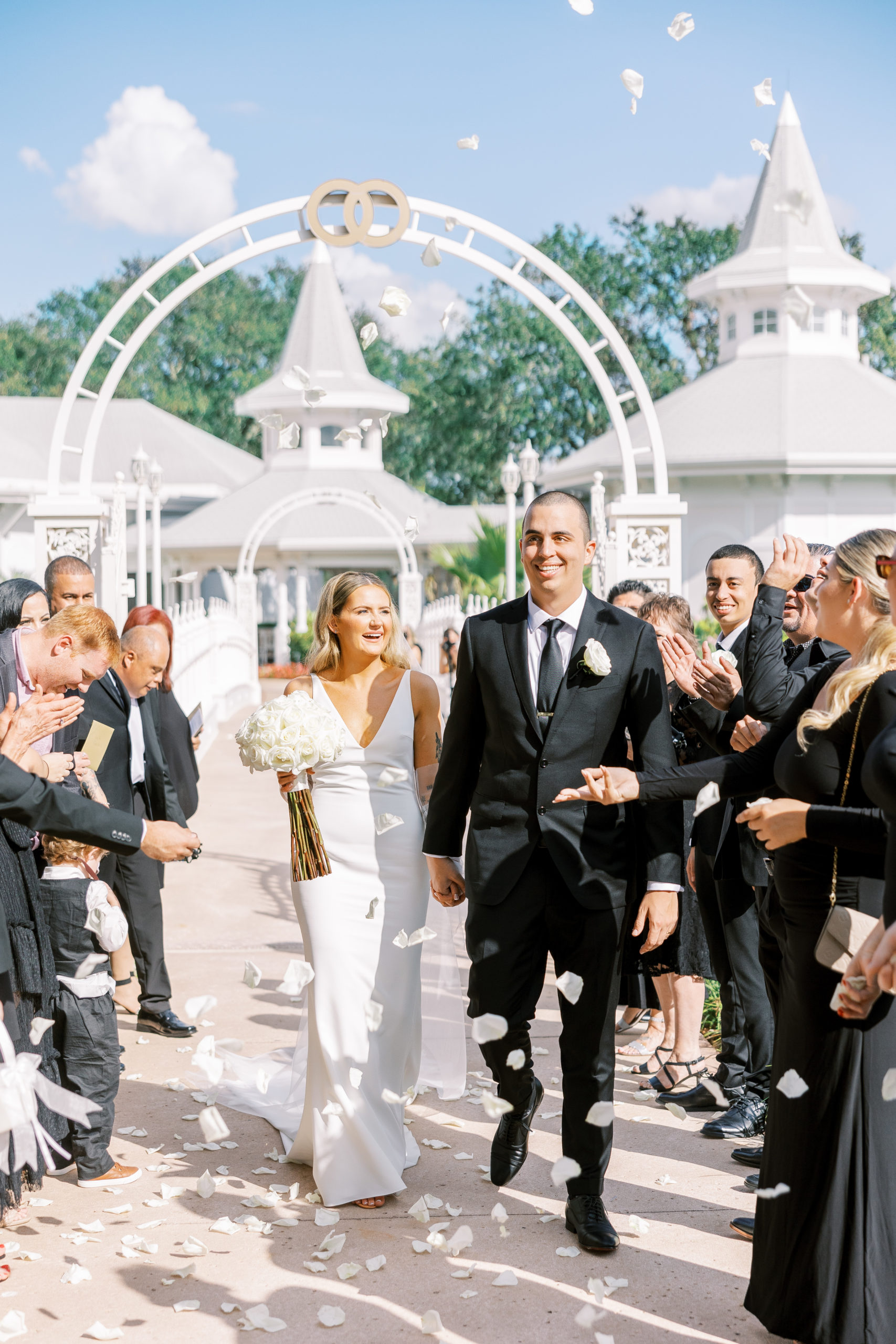 Bride and groom hold hands and smile as guests in black tie throw white petals on top of white bridge for Disney Film Wedding Photographer