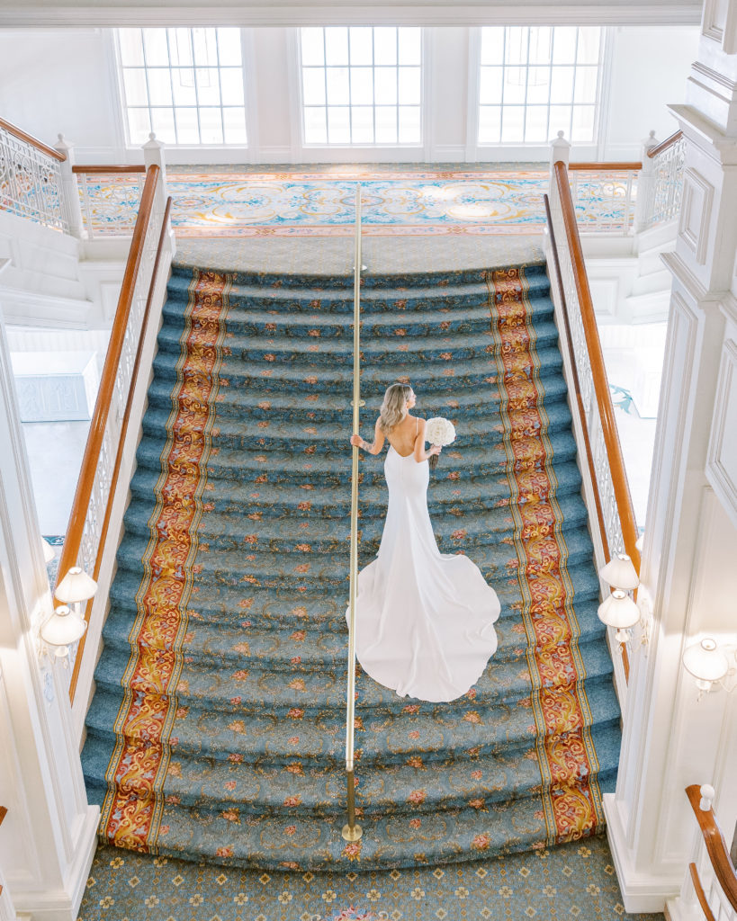 Aerial view of bride walking up grand staircase with blue and red carpet, holding white bouquet for Disney Film Wedding Photographer