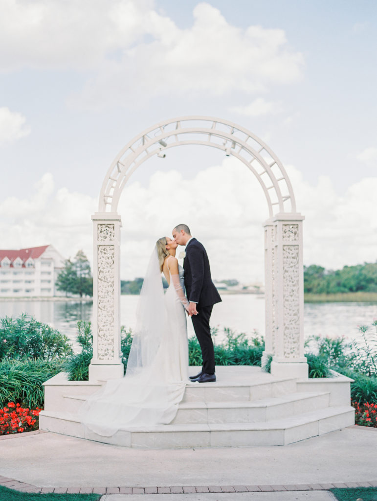 Bride and groom hold hands and kiss underneath white delicate archway with lake in background for California Grill Wedding