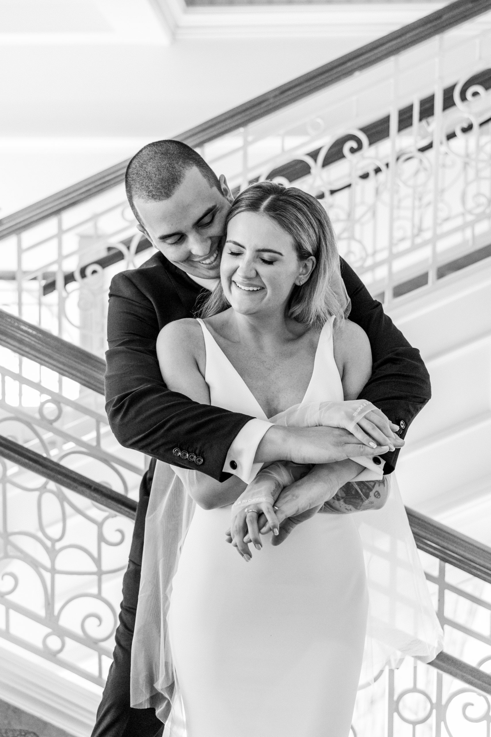 Groom wraps arms around bride from behind as they smile on grand staircase with delicate bannister for California Grill Wedding