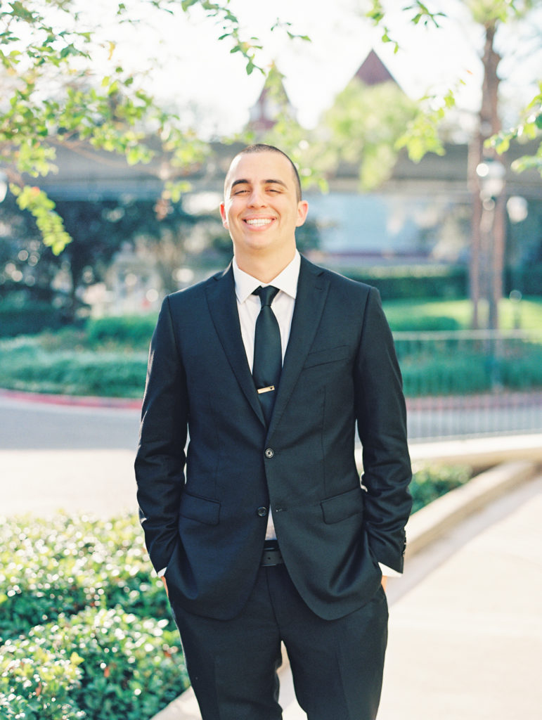 Groom smiles and poses with hands in pocket, wearing black suit 