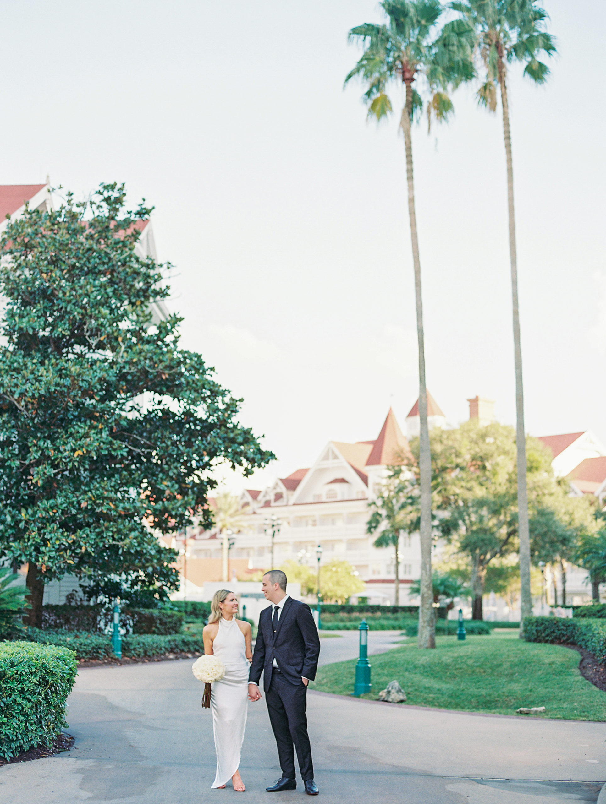 Bride and groom hold hands and smile on pathway with greenery and white and orange resort in background for California Grill Wedding