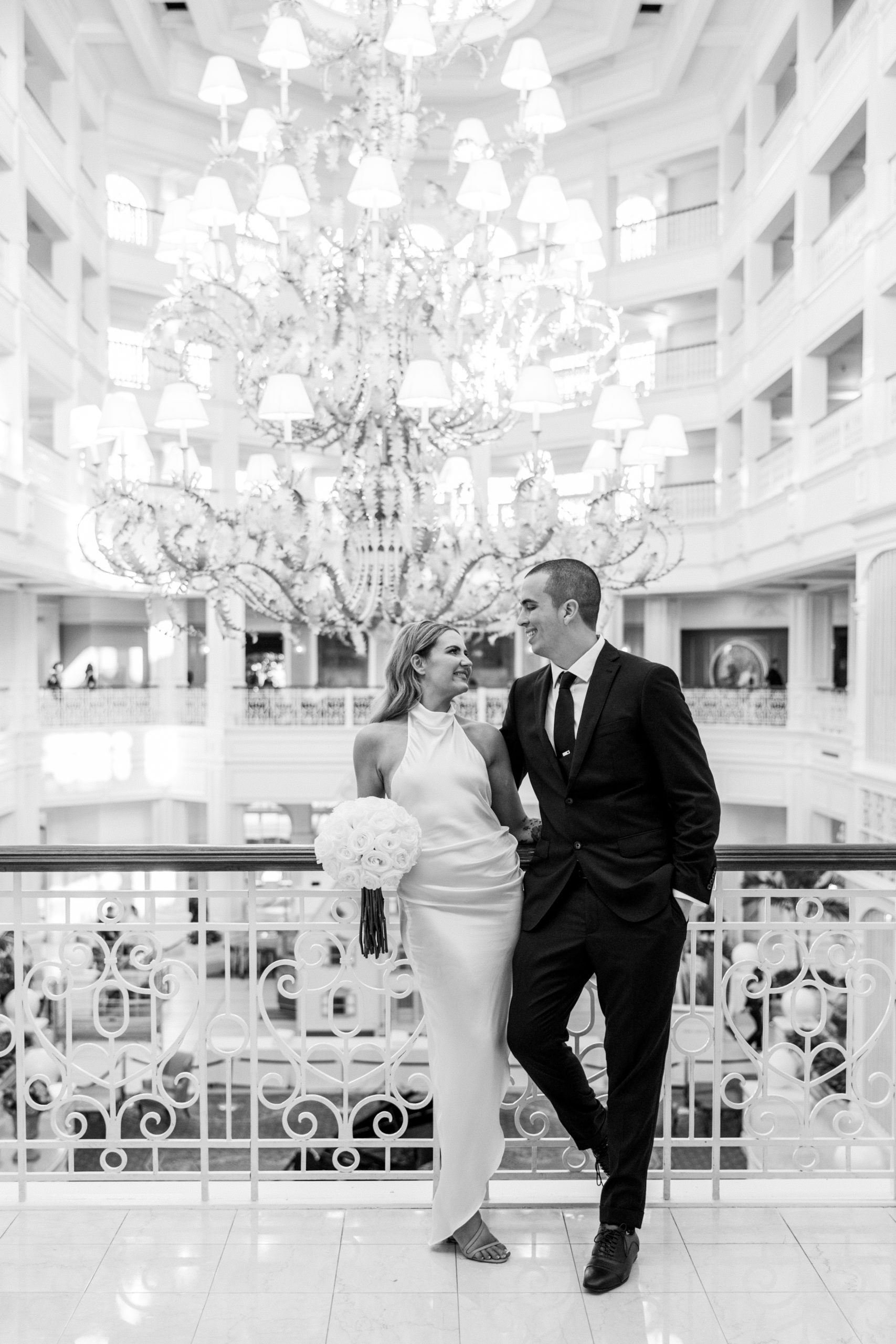 Black and white photo of bride and groom smiling against bannister and large chandelier behind them for Disney Film Wedding Photographer