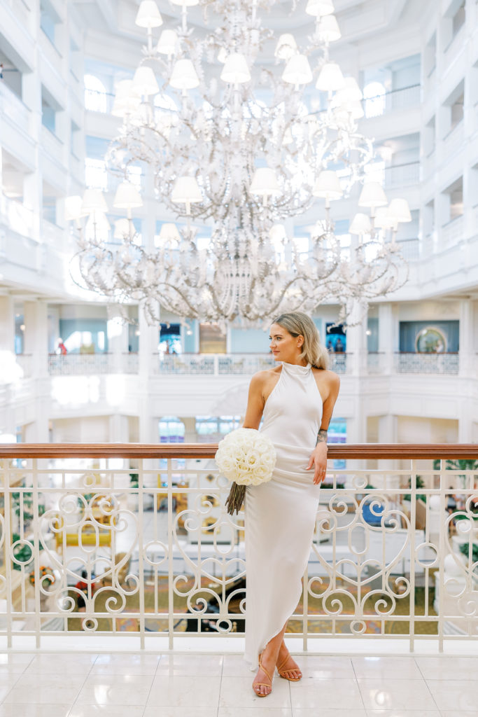 Bride rests against bannister and poses holding white rose bouquet with large silver chandelier for Disney Film Wedding Photographer