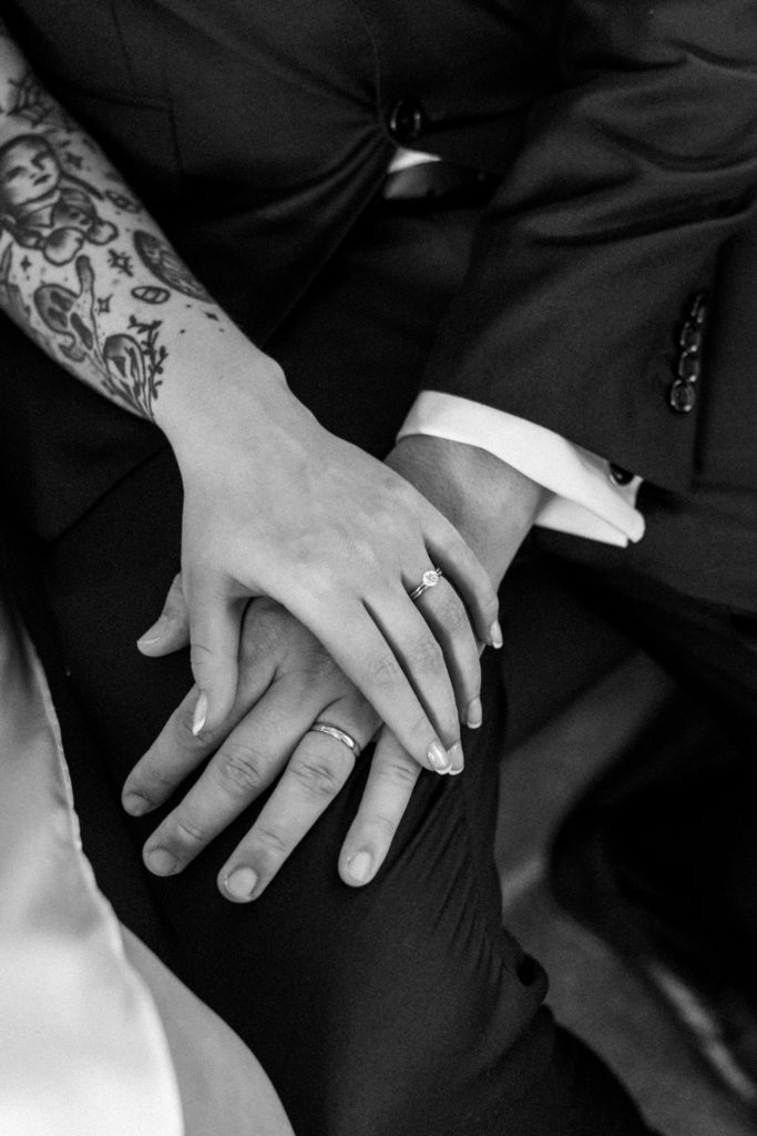 Black and white photo of bride and groom's hands resting on groom's knee, wearing wedding rings 