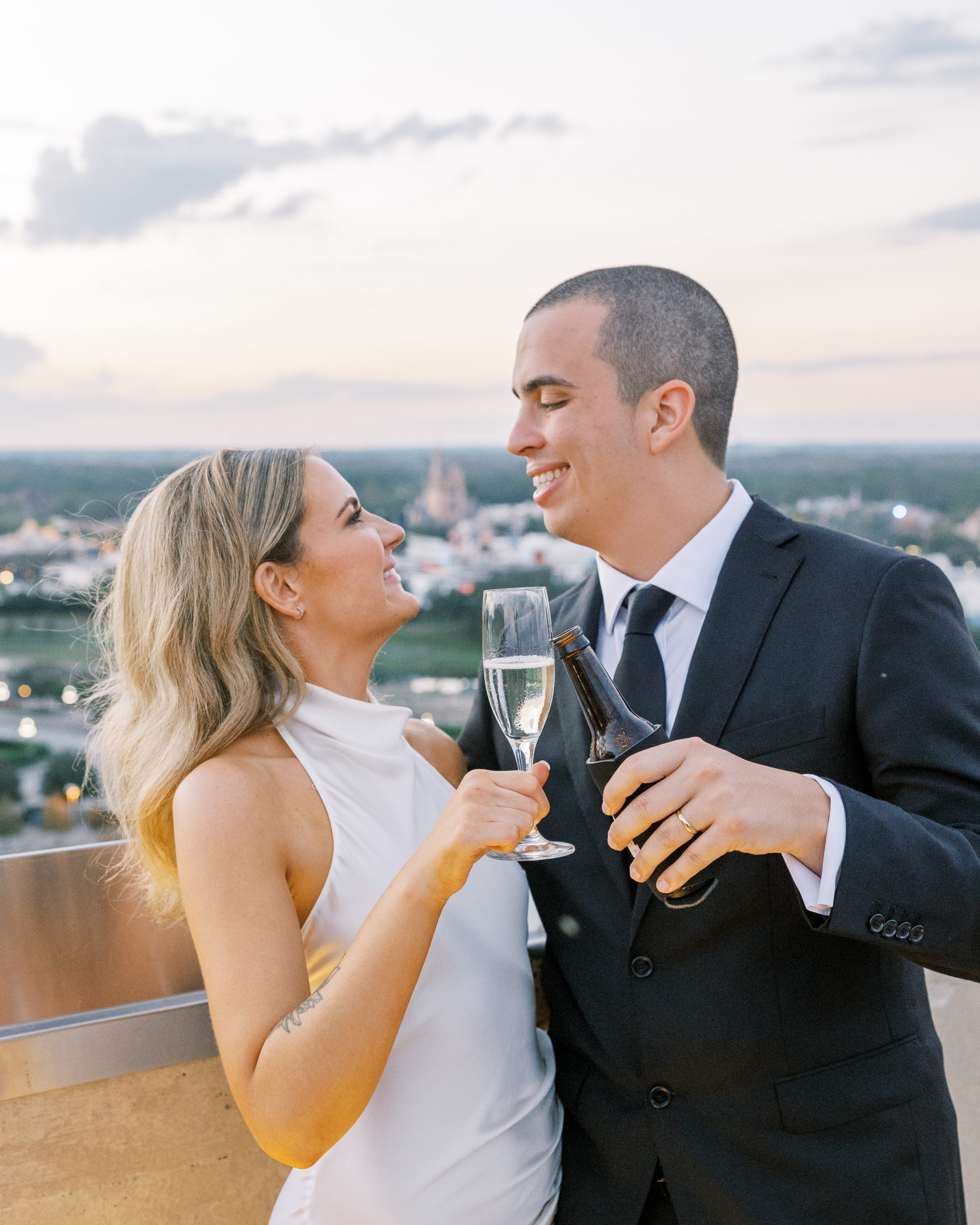Bride and groom smile at each other on porch with disney in the background, and do a cheers with champagne and beer for California Grill Wedding