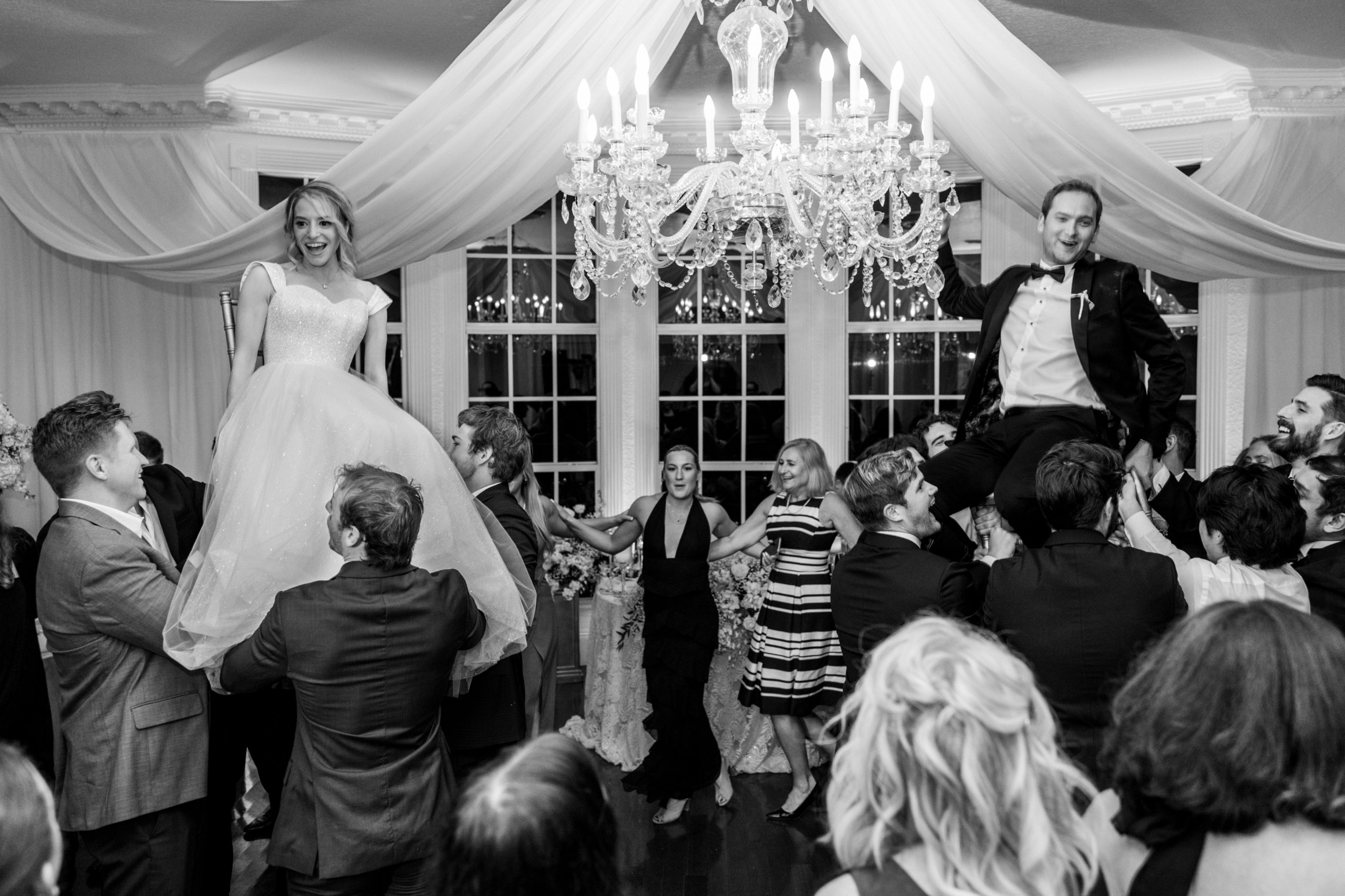 Bride and groom sit in chairs and are hoisted up on dance floor, known as the Hora, a traditional Jewish wedding tradition for Luxmore Grande Wedding
