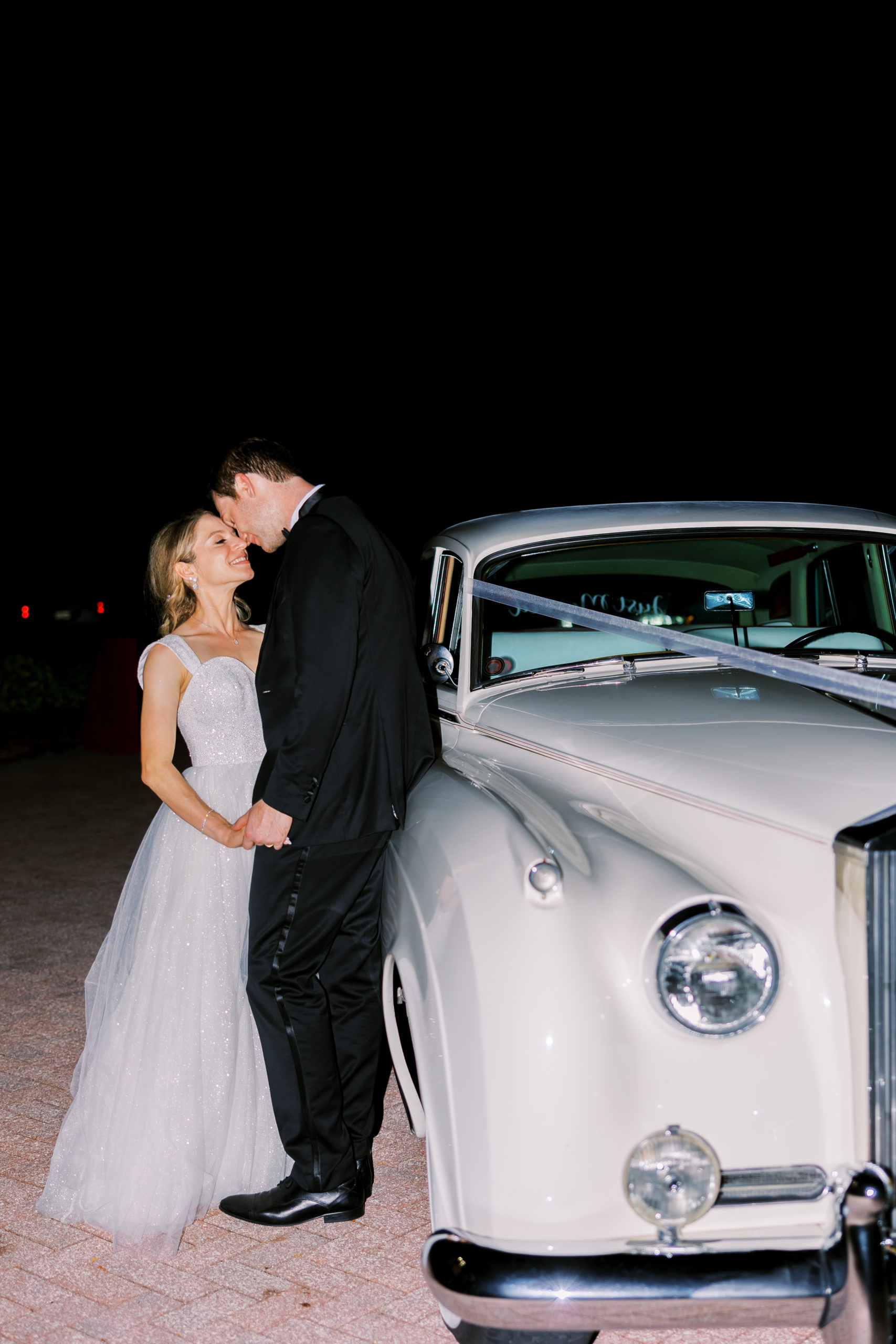 Bride and groom hold hands leaning in for a kiss against classic white car for Luxmore Grande Wedding