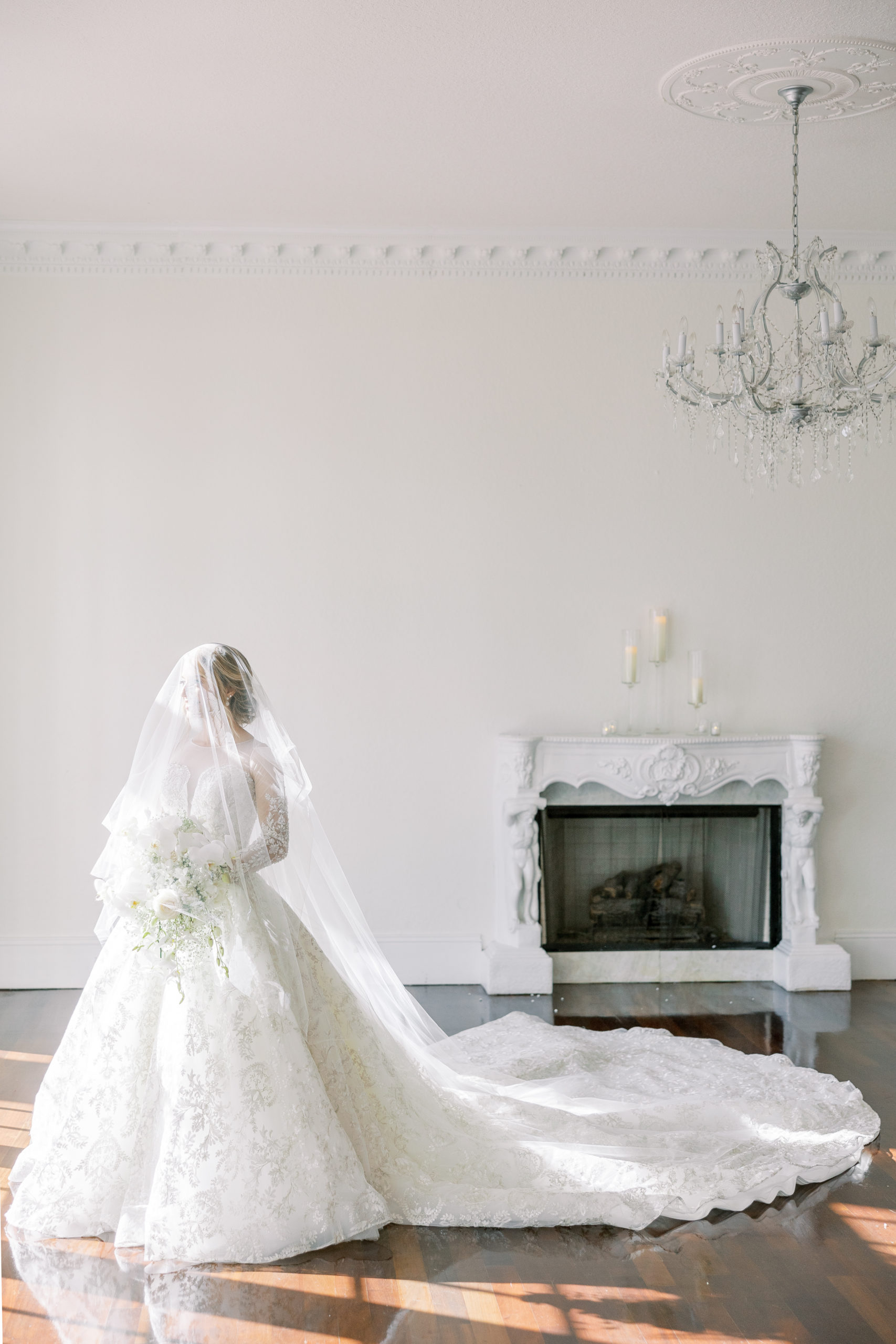 Bride poses looking away wearing veil over face with fireplace behind and chandelier above 