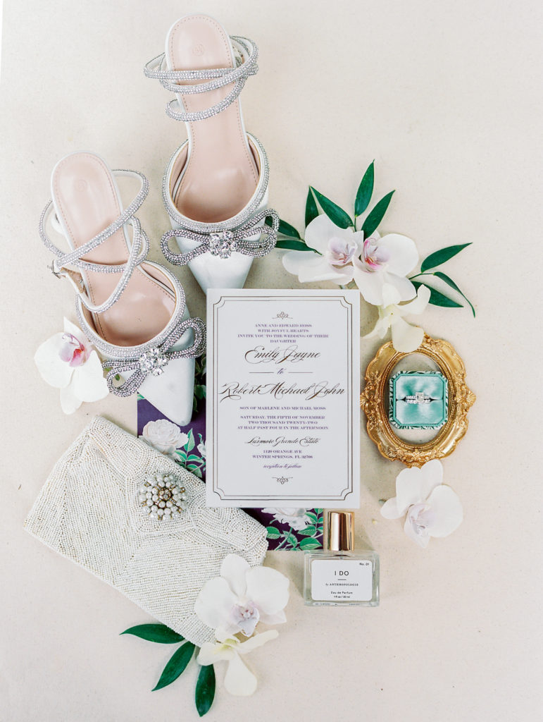 Flat lay of wedding heels, sparkled clutch, invitation, perfume, and rings in wedding box inside of gold frame