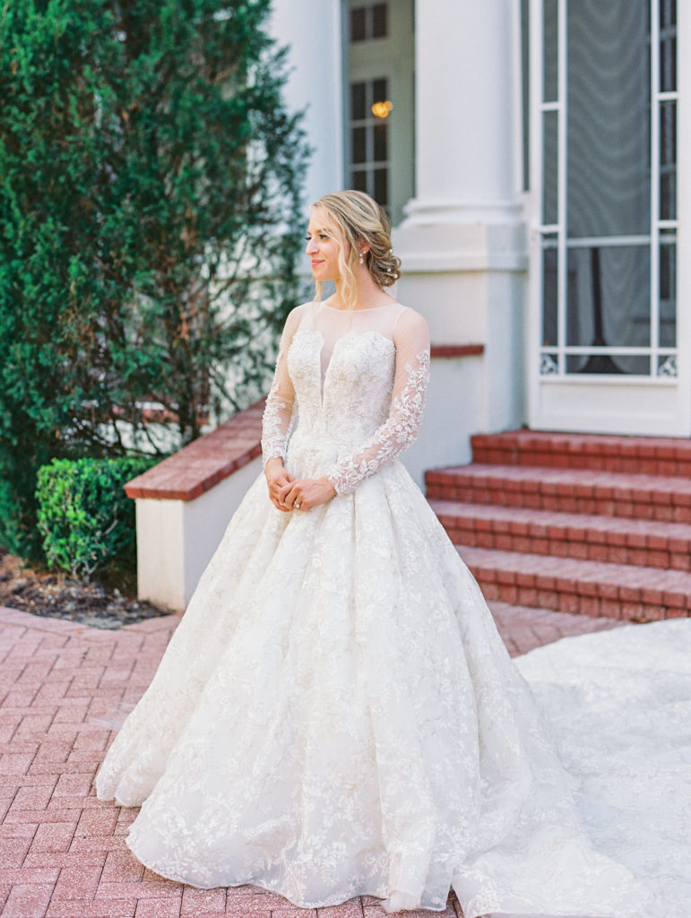 Bride poses holding her hands and looking away in front of brick stairs and white pillars 