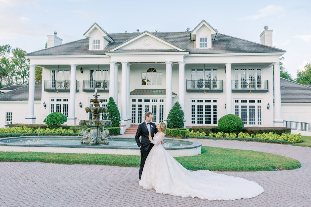 Bride places hand on groom's chest as they smile in front of grand estate and fountain 