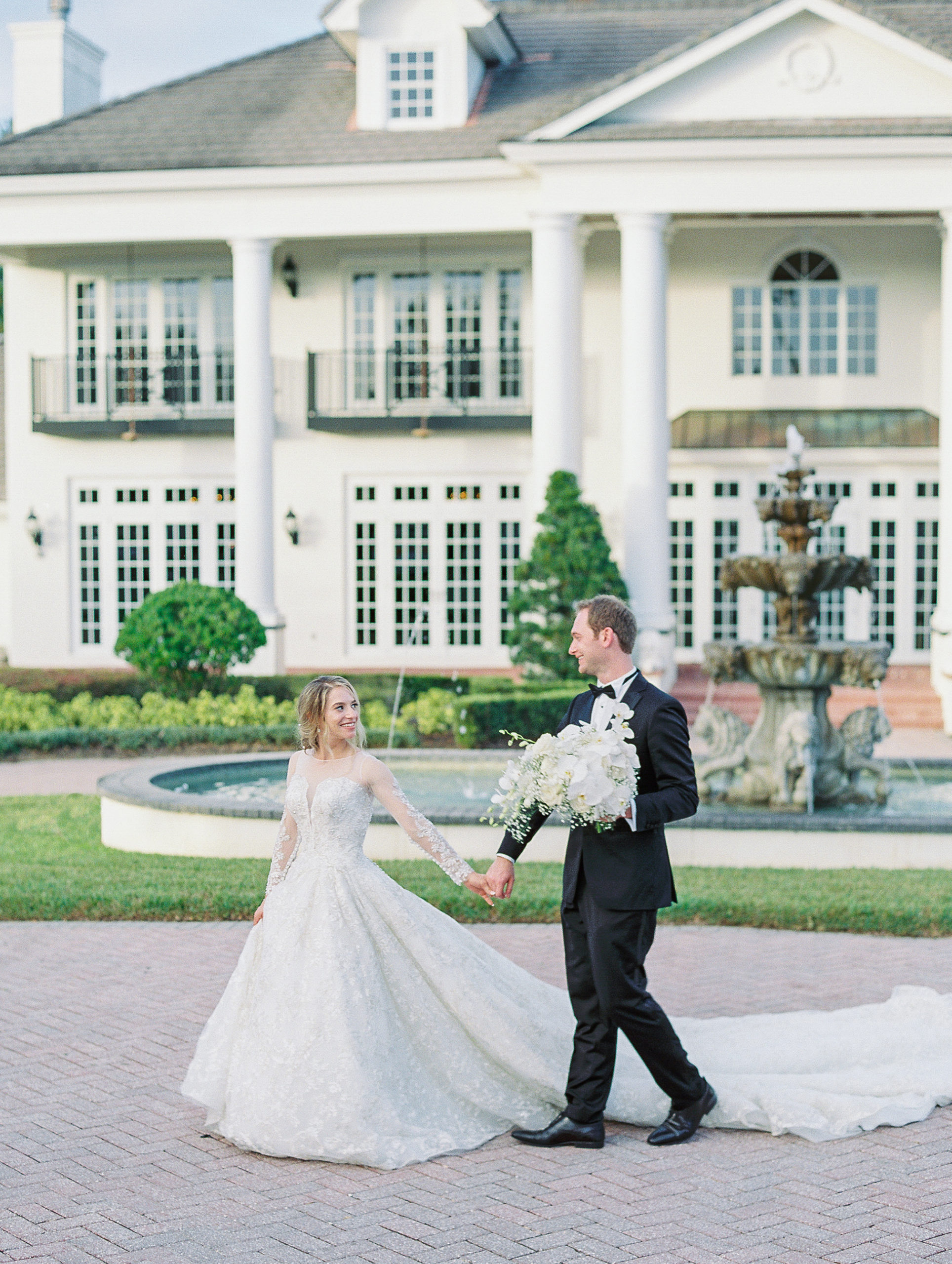 Bride leads groom across brick driveway in front of large estate and fountain for Luxmore Grande Wedding