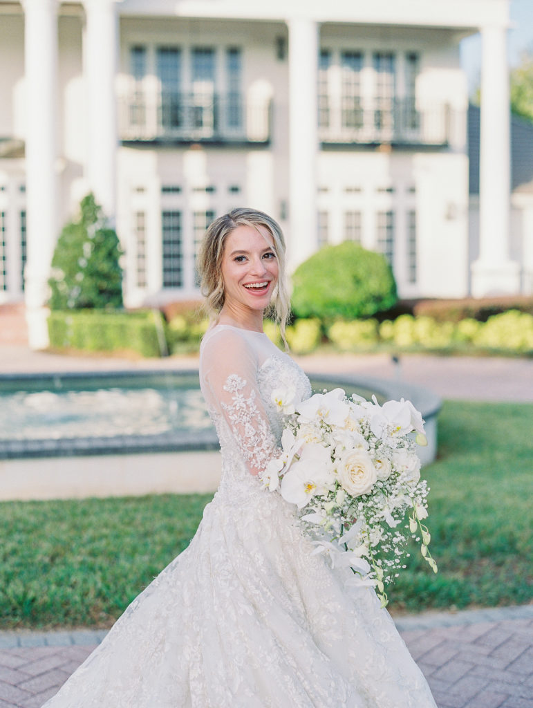Bride smiles and looks over shoulder holding white bouquet in front of white estate and fountain for Luxmore Grande Wedding
