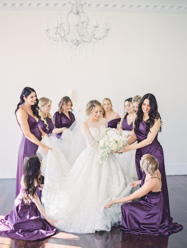 Bridesmaids surround bride wearing purple dresses and help adjust wedding dress 