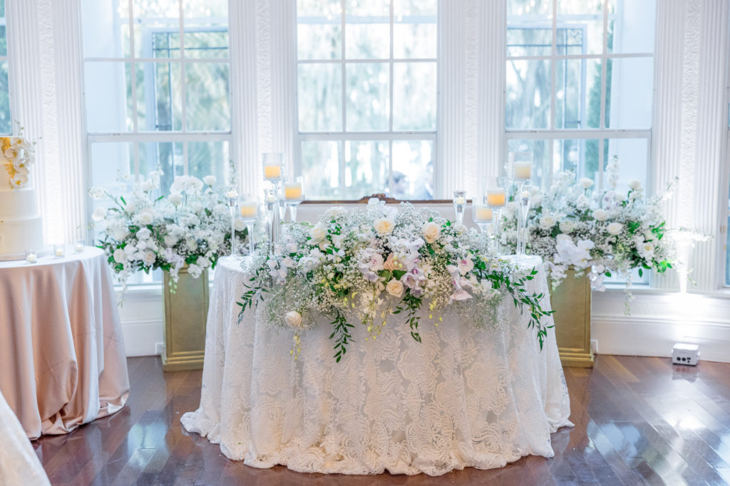 Bride and groom's reception table with white tablecloth and elegant white rose arrangements against windows for Luxmore Grande Wedding
