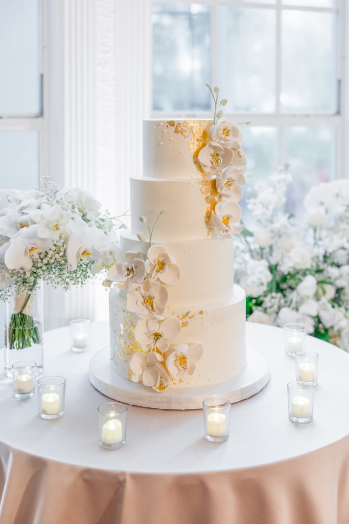 4-tier white wedding cake with gold flakes and white flowers surrounded by small tea light candles 