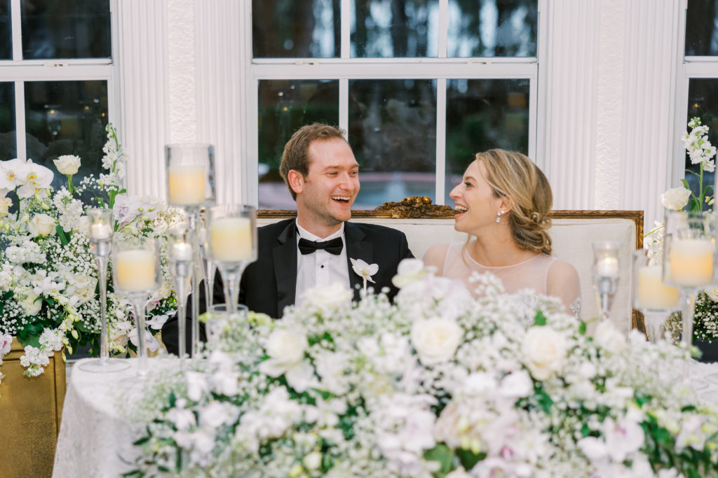 Bride and groom sit at personal table and laugh with white flower arrangements and candles for Luxmore Grande Wedding