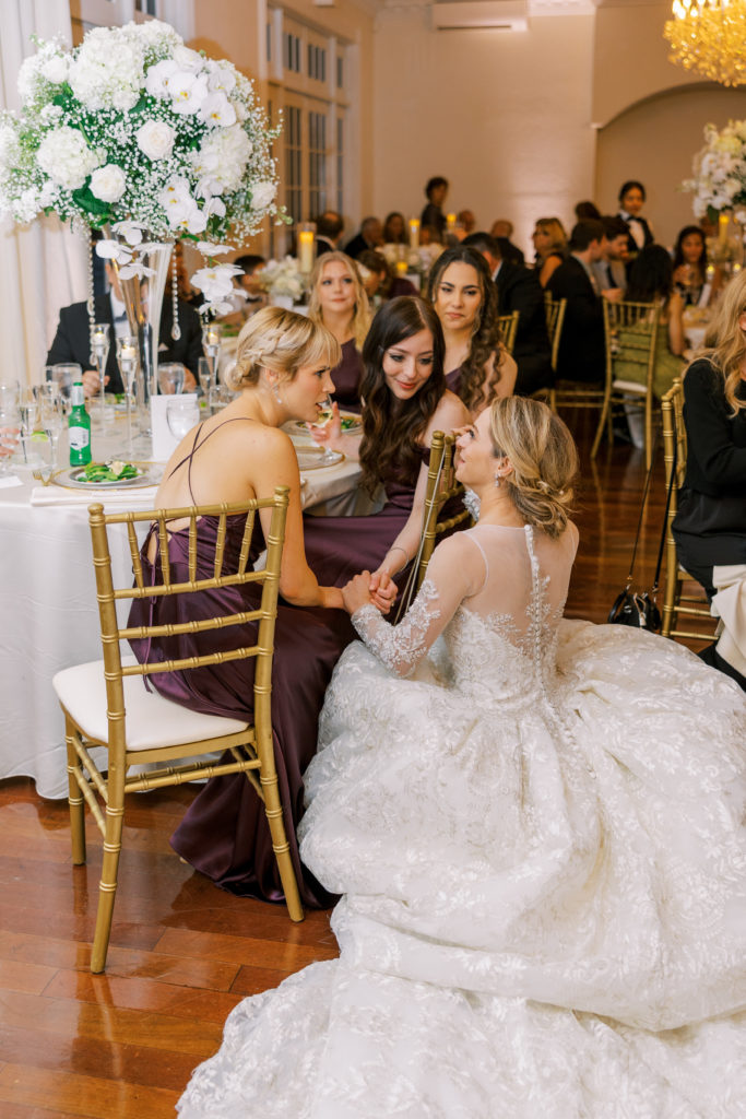 Bride kneels down to talk to bridesmaids during wedding reception 
