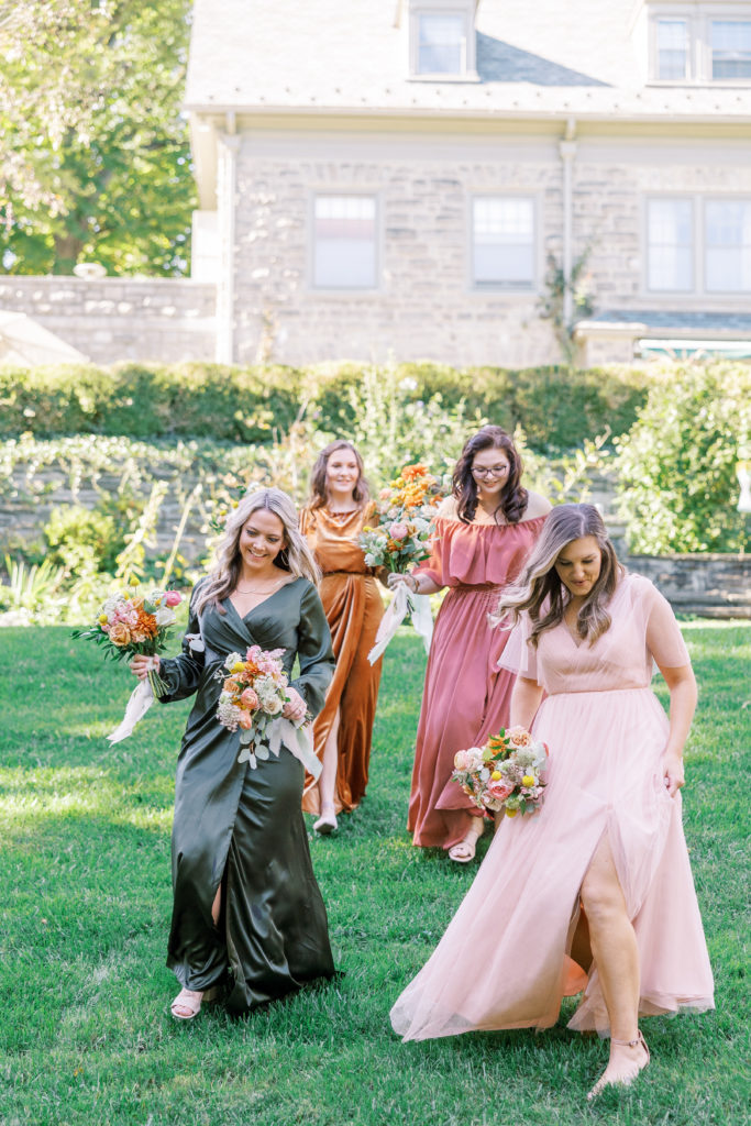Bridesmaids hold bouquets and walk across lawn wearing different colored fall dresses of pink, orange, and green 