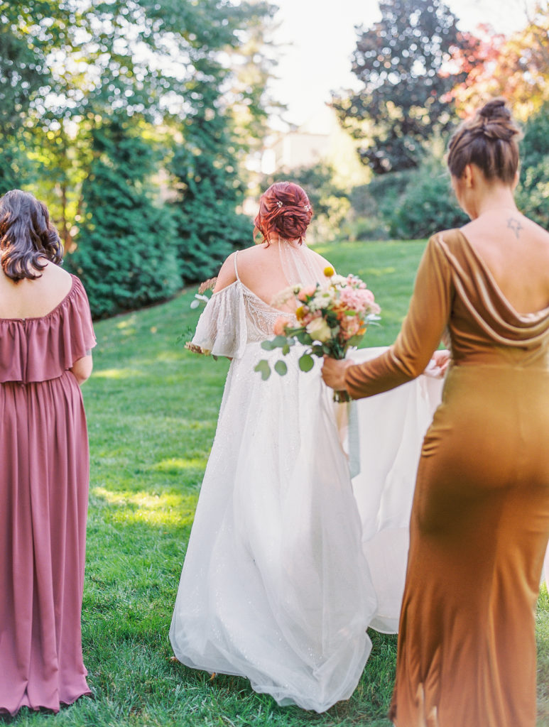 Bride walks across lawn with bridesmaids as they hold dress up 
