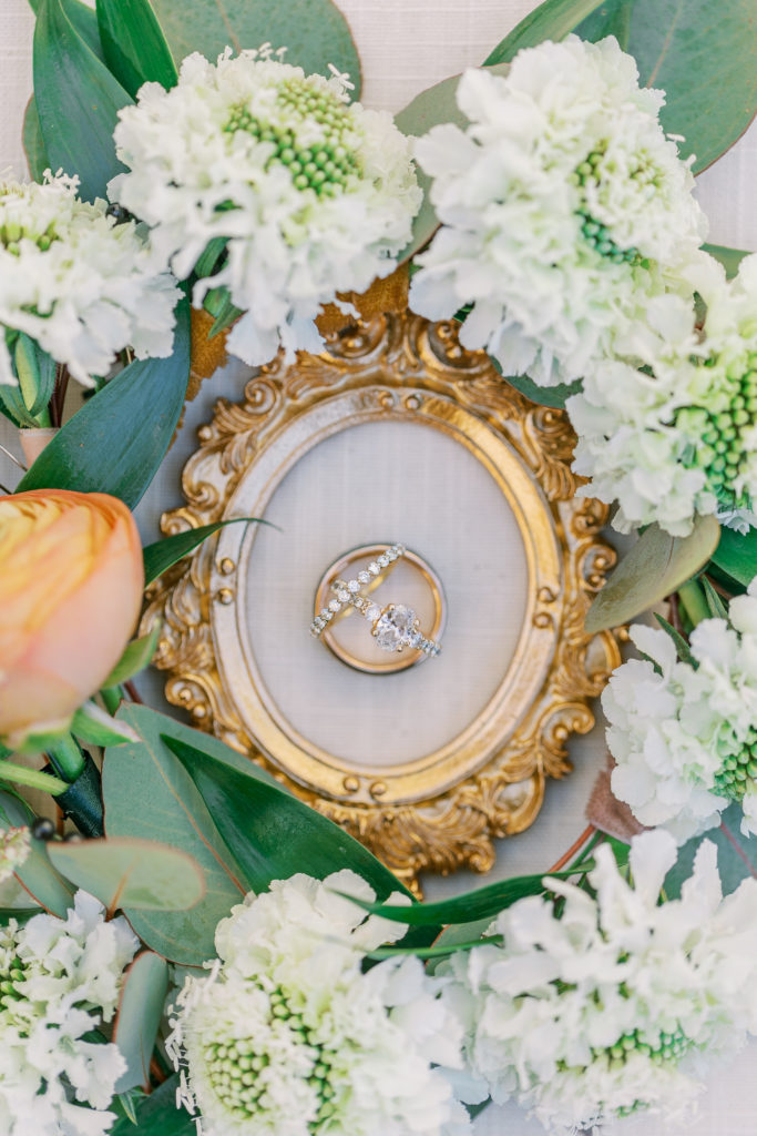 Wedding rings inside of gold frame and white and peach flowers 