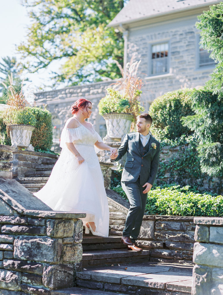 Groom leads bride down stone stairs out of garden wearing a grey suit for philadelphia wedding photographer