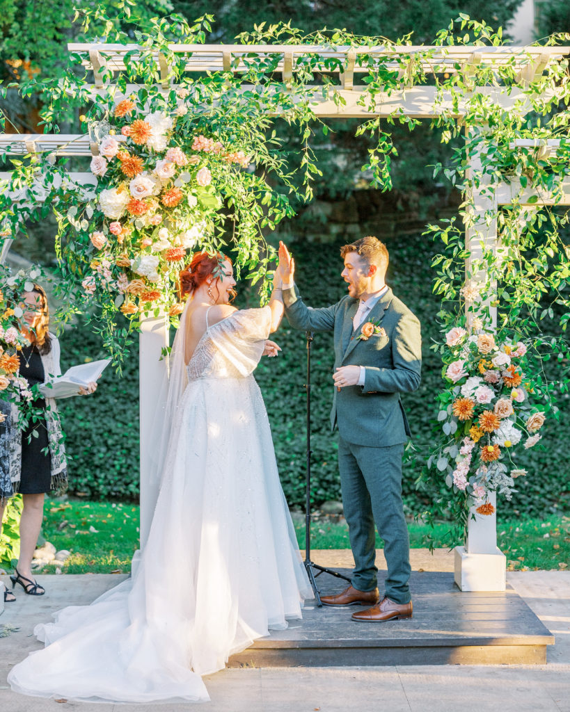 Bride and groom high five at the altar with large flower arrangements and white arch for philadelphia wedding photographer