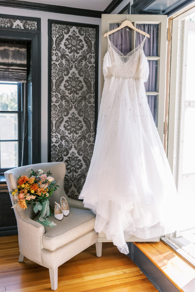 Wedding dress hangs upon door frame with bouquet and flowers on chair 